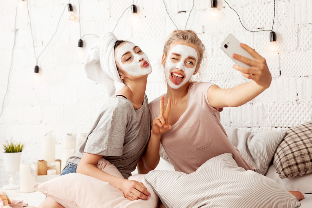 two girls wearing face masks take a selfie on a bed