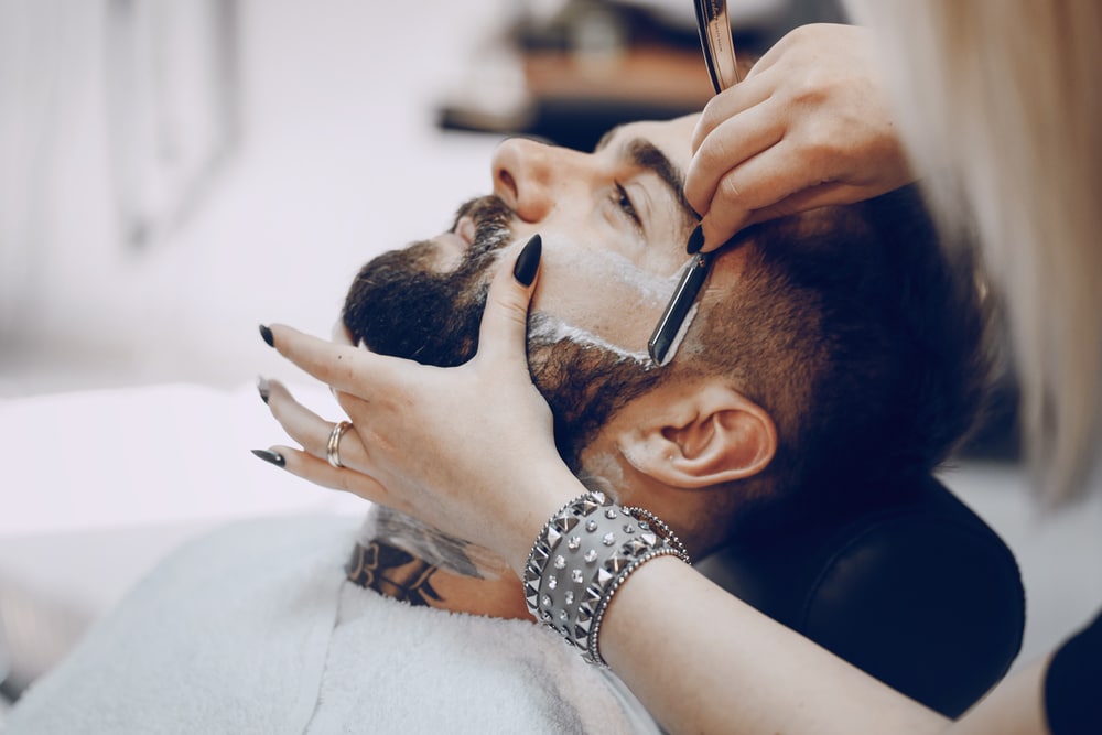 Female barber shaving male client's beard with straight razor